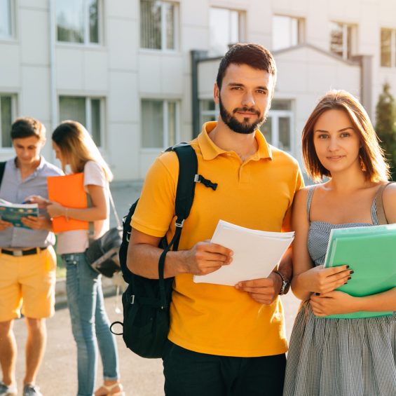 Two happy students with backpacks and books in their hands smiling at camera while standing on background of university and friends Two happy students with backpacks and books in their hands smiling at camera while standing on background of university and friends
